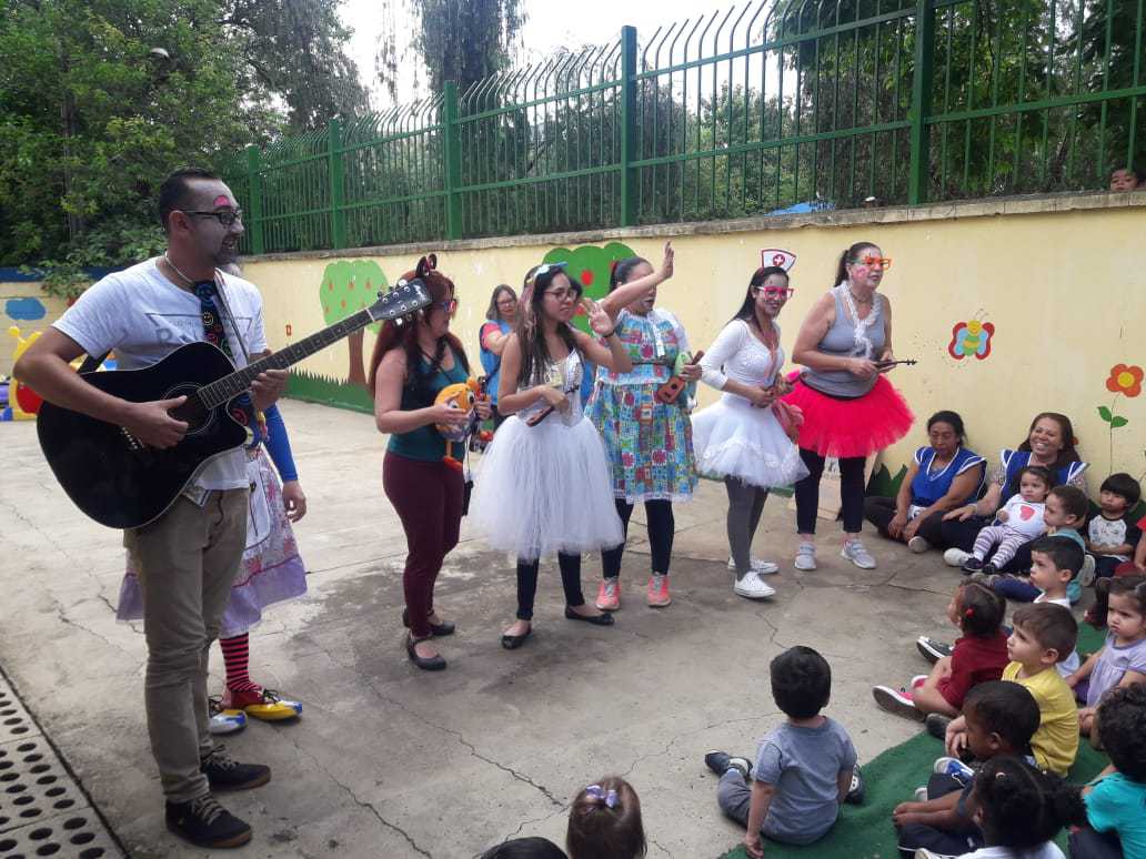Na imagem, Glorinha aparece com outros colegas de trabalho em uma apresentação para crianças. Há um homem tocando violão e mais seis mulheres caracterizadas com elementos infantis entretendo as crianças